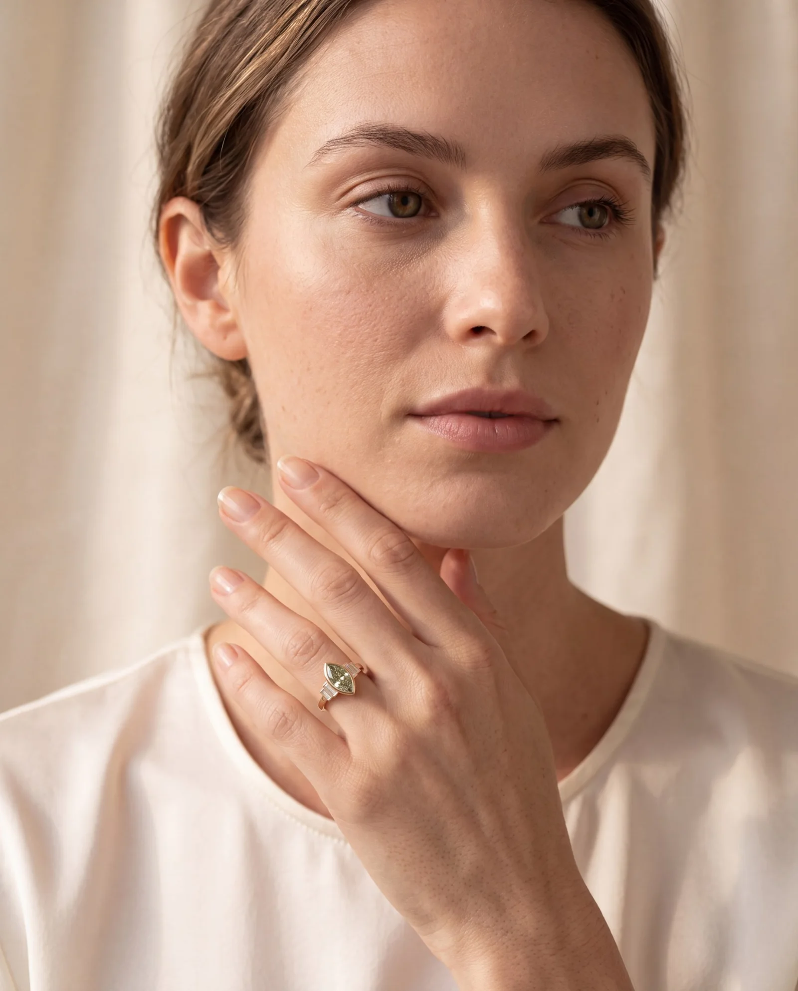 A woman touches her jaw, wearing a marquise lab-grown diamond ring in a gold bezel with baguette side stones